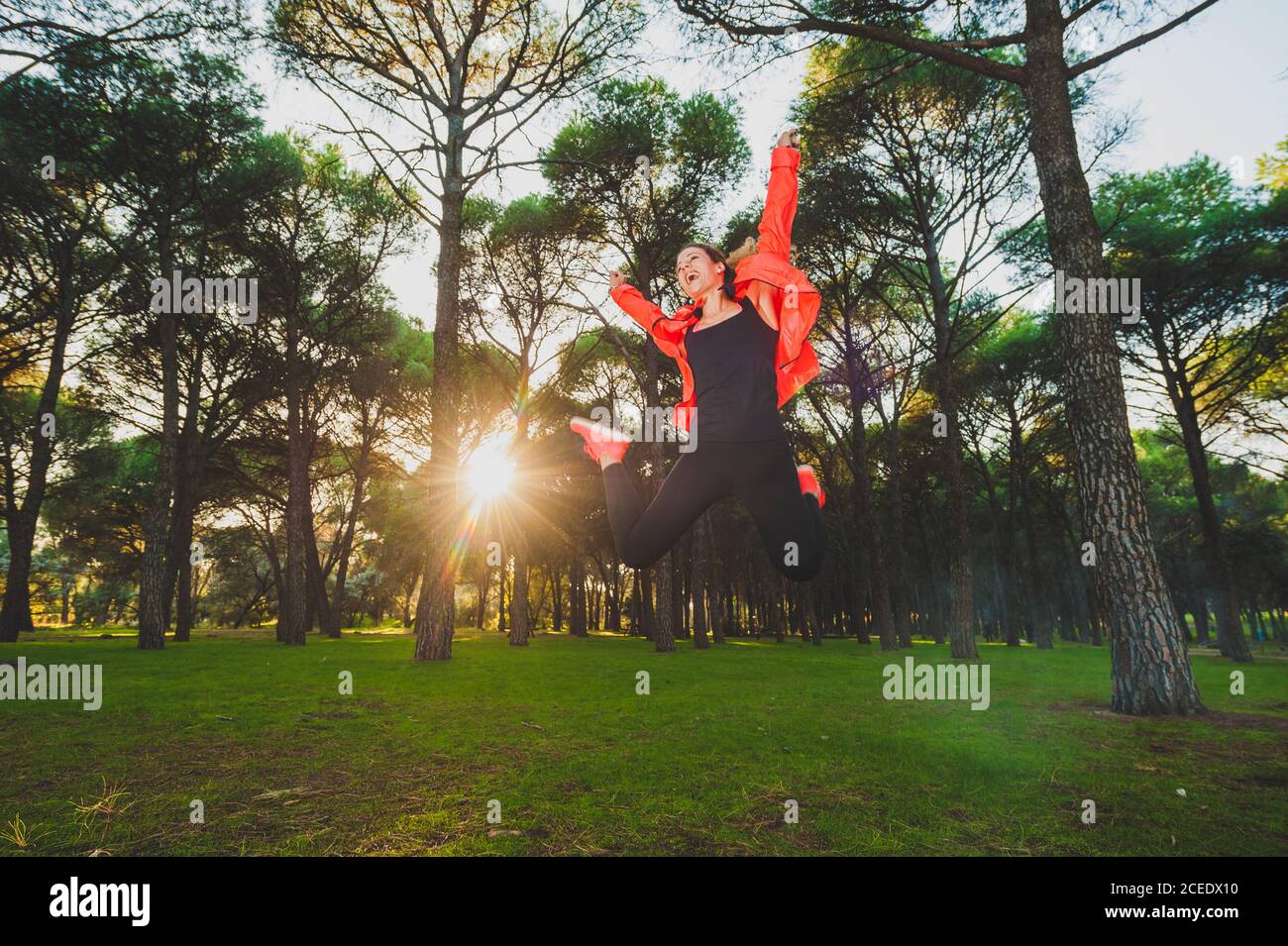 Bright happy Woman in sportive outfit jumping high above green meadow in sunny park Stock Photo
