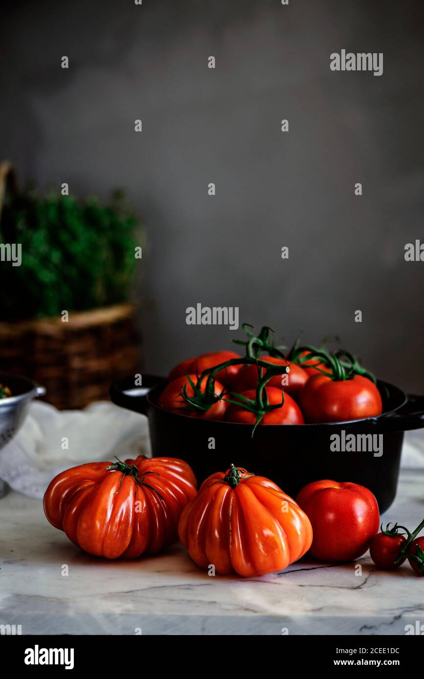 Big red ripe tomatoes of different forms in pot on table near grey wall Stock Photo