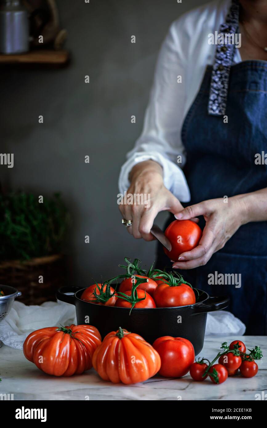 Crop lady with knife cutting vegetable near pot with red fresh tomatoes on table Stock Photo