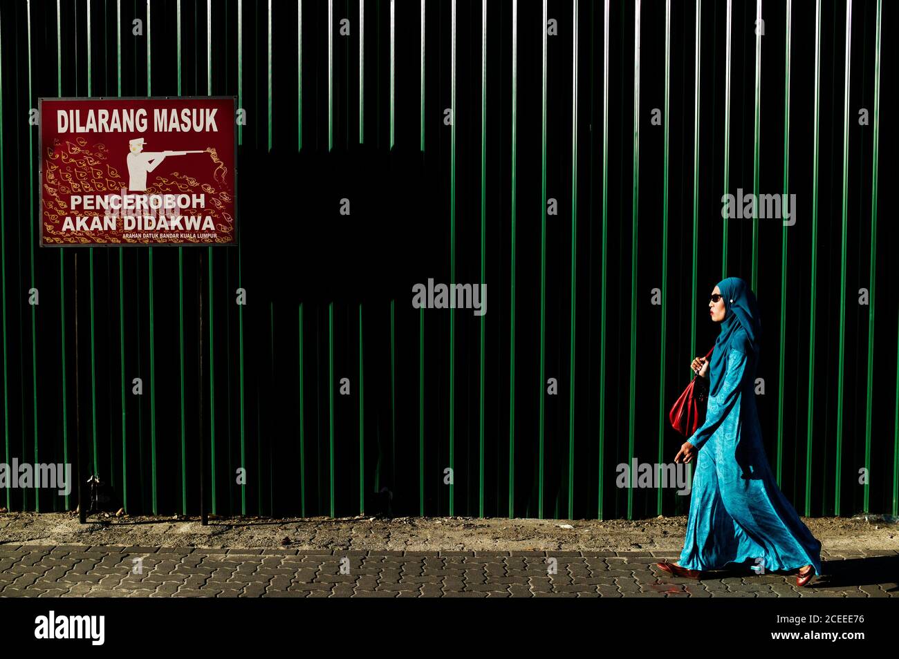Side view of Woman wearing blue abaya and headscarf walking on street on background of metal fence. Stock Photo