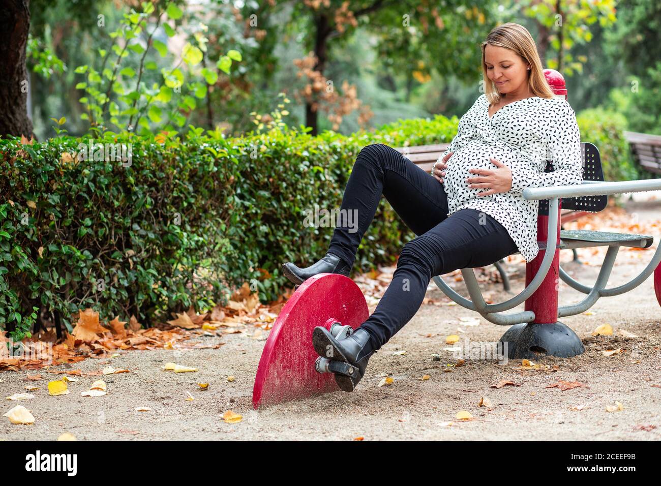 Pregnant smiling lady holding belly and sitting at toy cycle on playground near green bushes Stock Photo