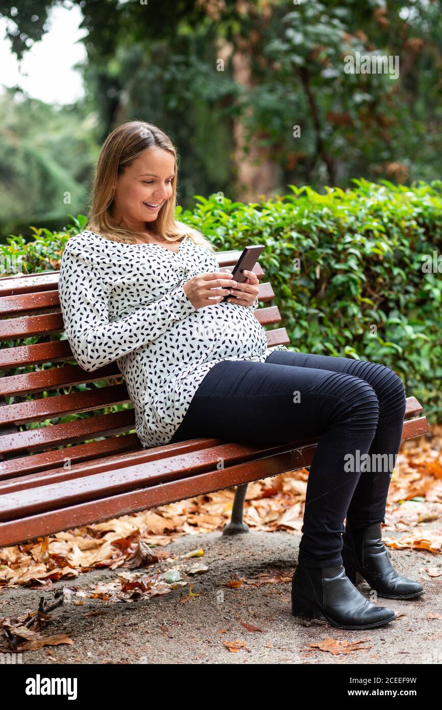 Pregnant smiling lady using mobile phone and sitting on seat in park Stock Photo