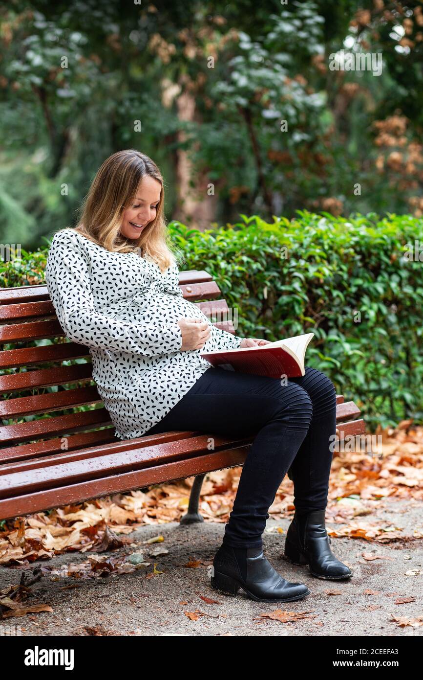 Pregnant smiling lady holding belly and reading volume on seat in park Stock Photo