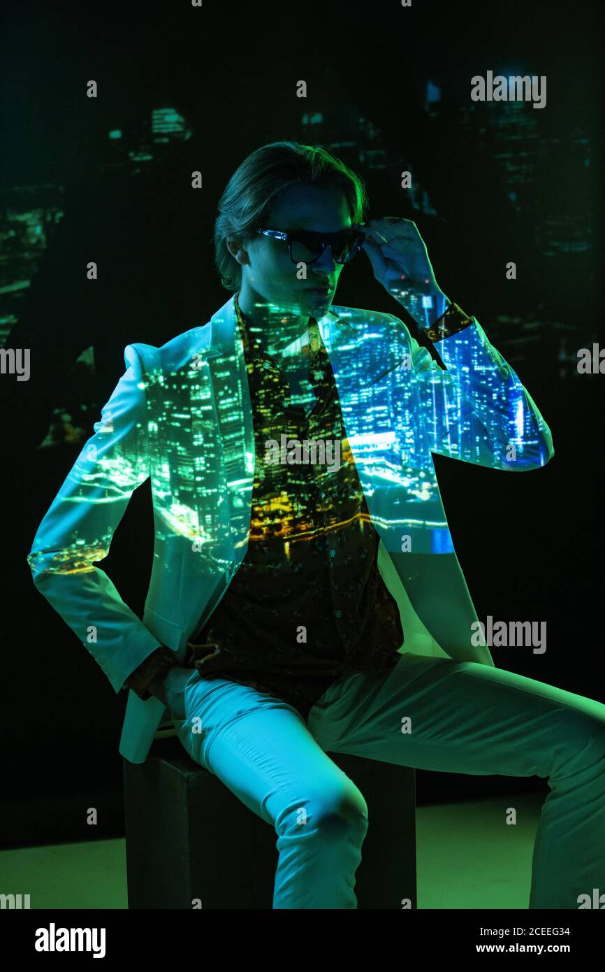 Picture of modern city projected on androgynous male model in sunglasses and suit sitting on cube in dark room Stock Photo