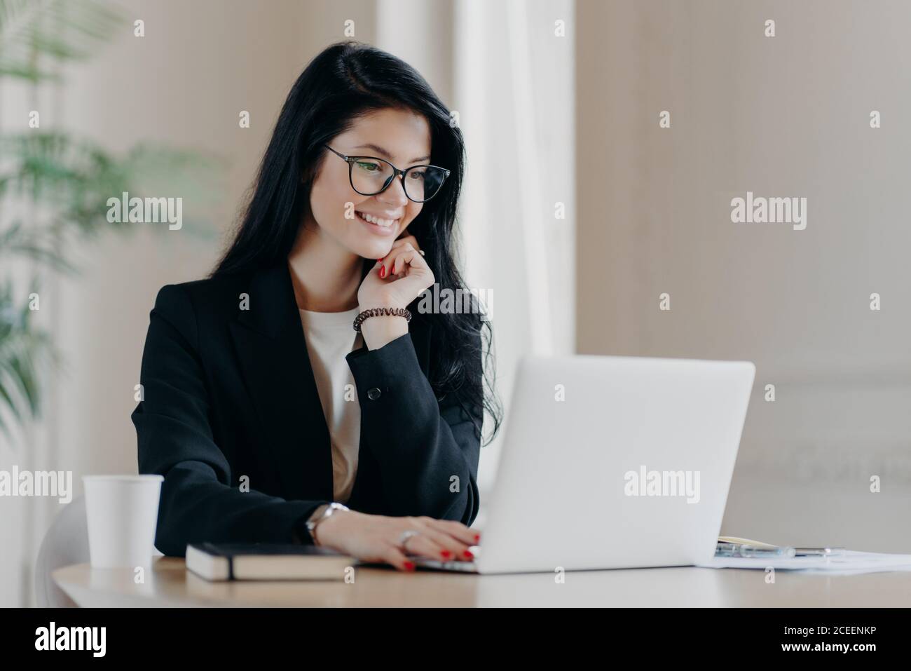 Cheerful female employee types on laptop computer, works remotely, concentrated with smile at device screen, writes email letter, works in office drin Stock Photo