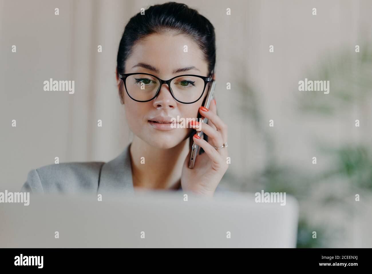 Horizontal shot of dark haired experienced woman with serious expression, makeup, talks via mobile phone, concentrated into screen, calls business par Stock Photo