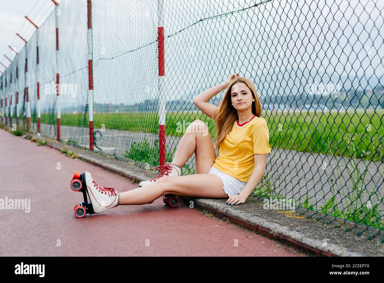 Pretty cheerful teen girl wearing shorts with roller skates and sitting on ground smiling at