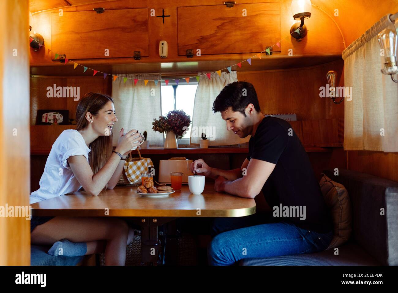 Side view of young man and Woman smiling and having good breakfast while sitting at table inside cozy caravan Stock Photo