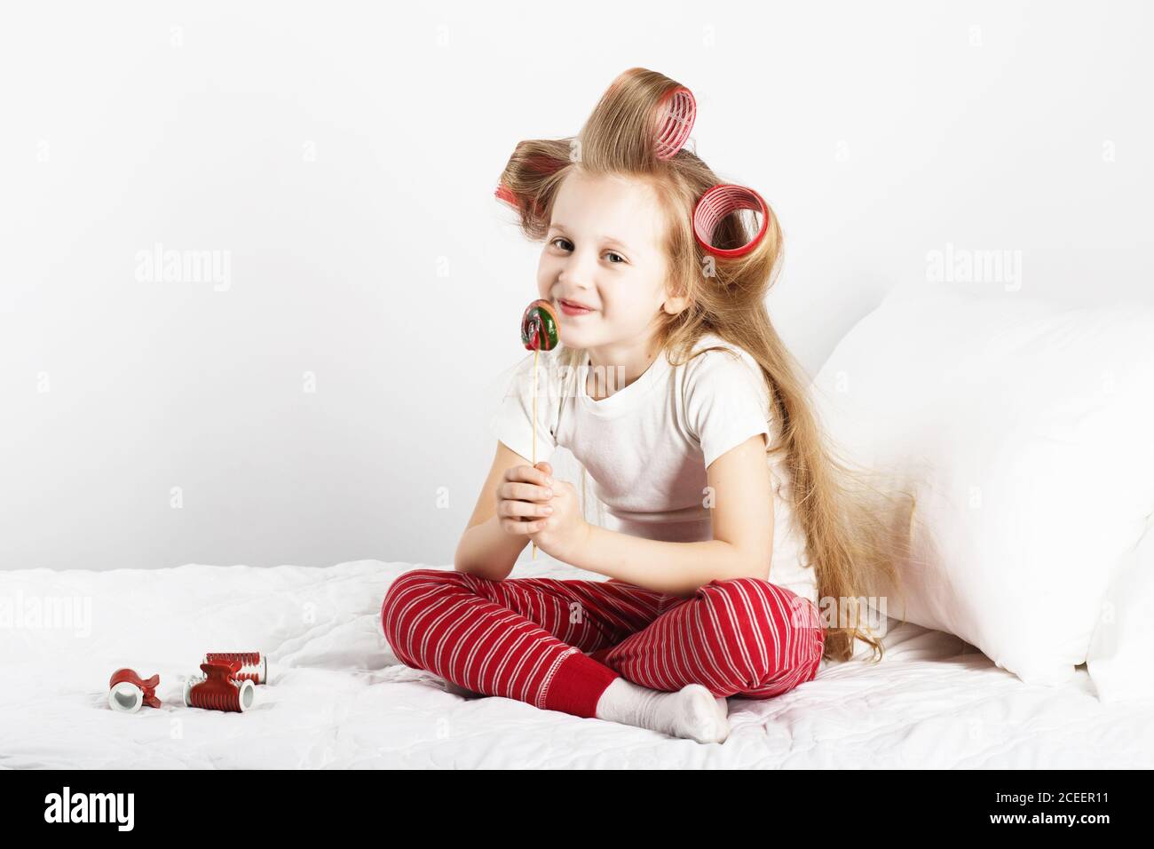 Portrait of a lovely little girl with big curlers on her head eating delicious lollipop against a white background. Day off, holiday, day of rest, fre Stock Photo