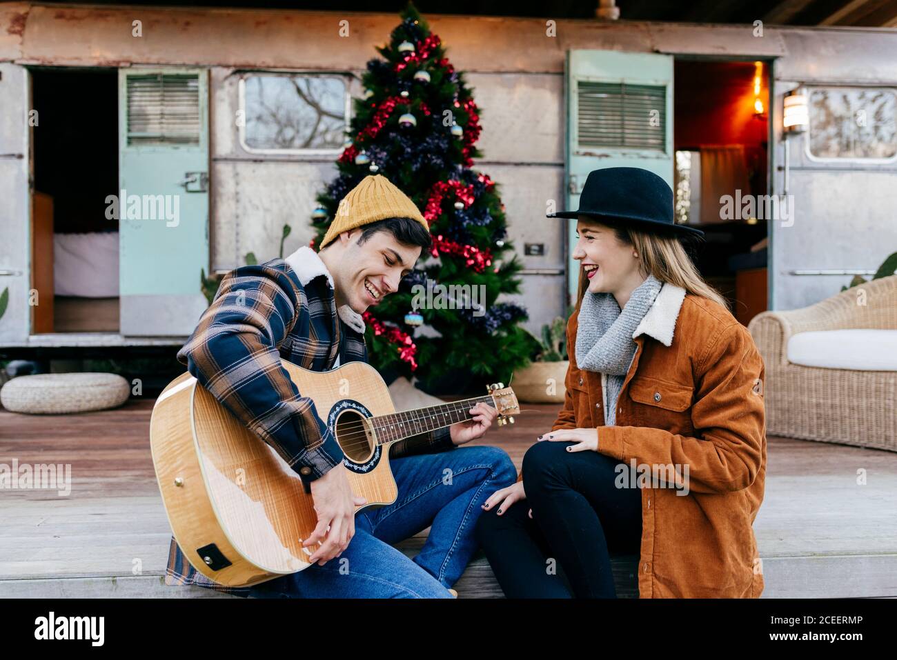 Handsome young man smiling and playing guitar for lovely girlfriend while sitting near Christmas tree and weathered trailer Stock Photo