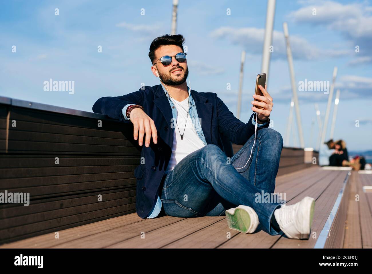 Trendy dandy man in sunglasses and jacket chilling with smartphone and headphones on promenade. Stock Photo