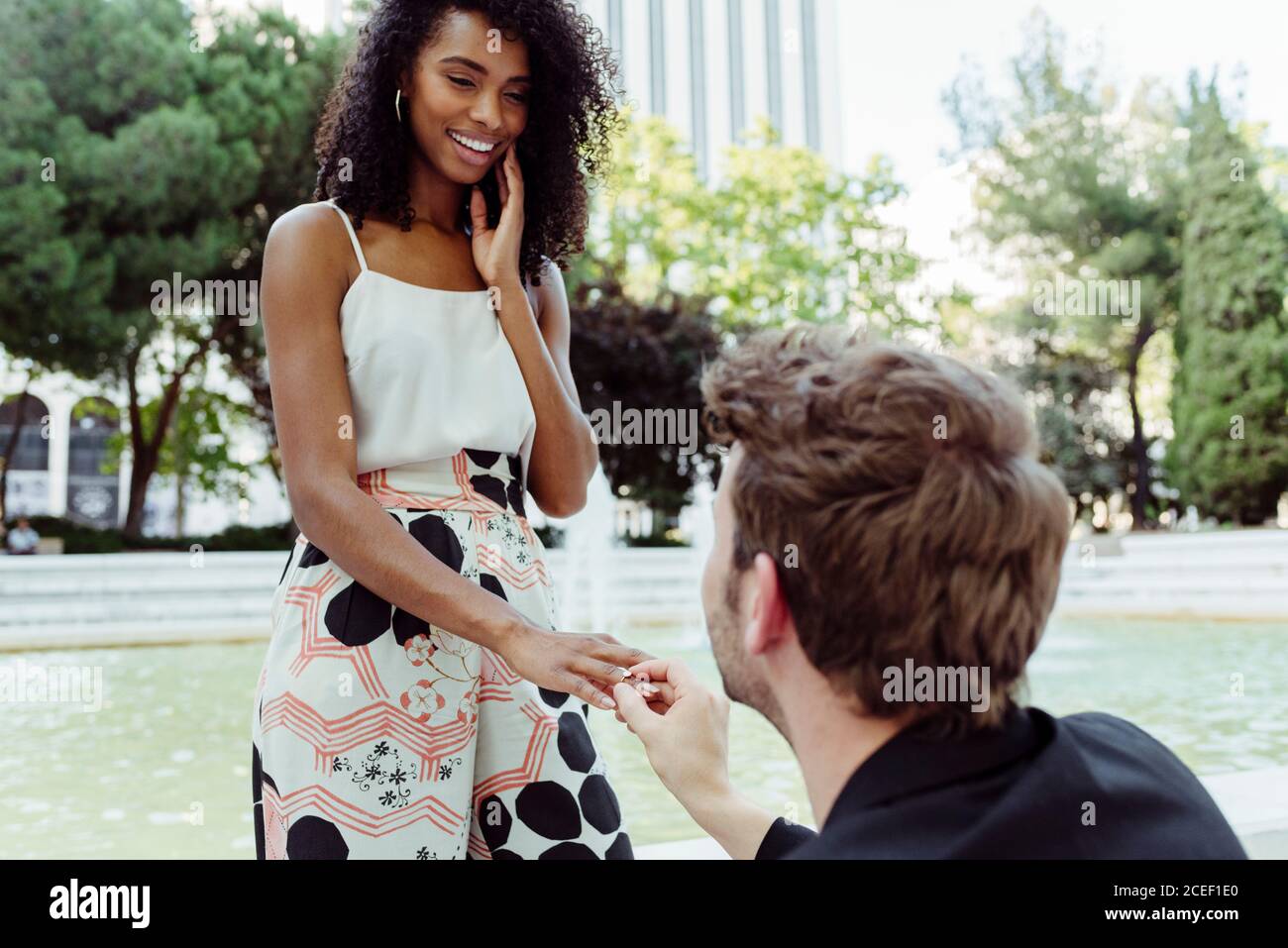 Caucasian man putting engagement ring on finger of black Woman while proposing to her near fountain in park Stock Photo