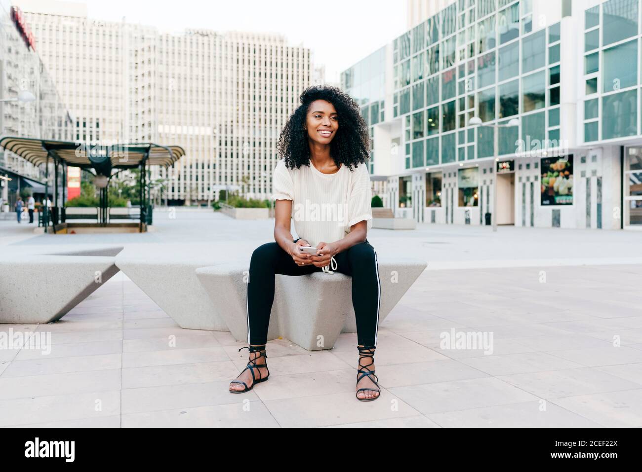 Pretty African-American lady smiling and looking at camera while sitting on city street and browsing modern smartphone Stock Photo