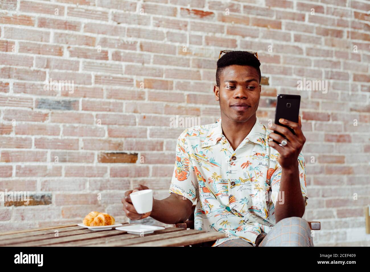 Handsome black man drinking hot beverage and browsing smartphone while sitting at small table near brick wall Stock Photo