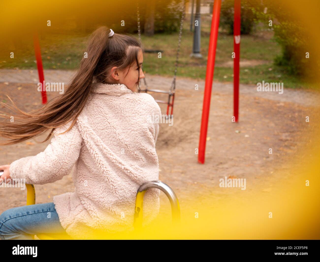View through railings over happy smiling teenage girl in a faux fur coat and with long ponytail spinning at a merry-go-round at children playground. Stock Photo