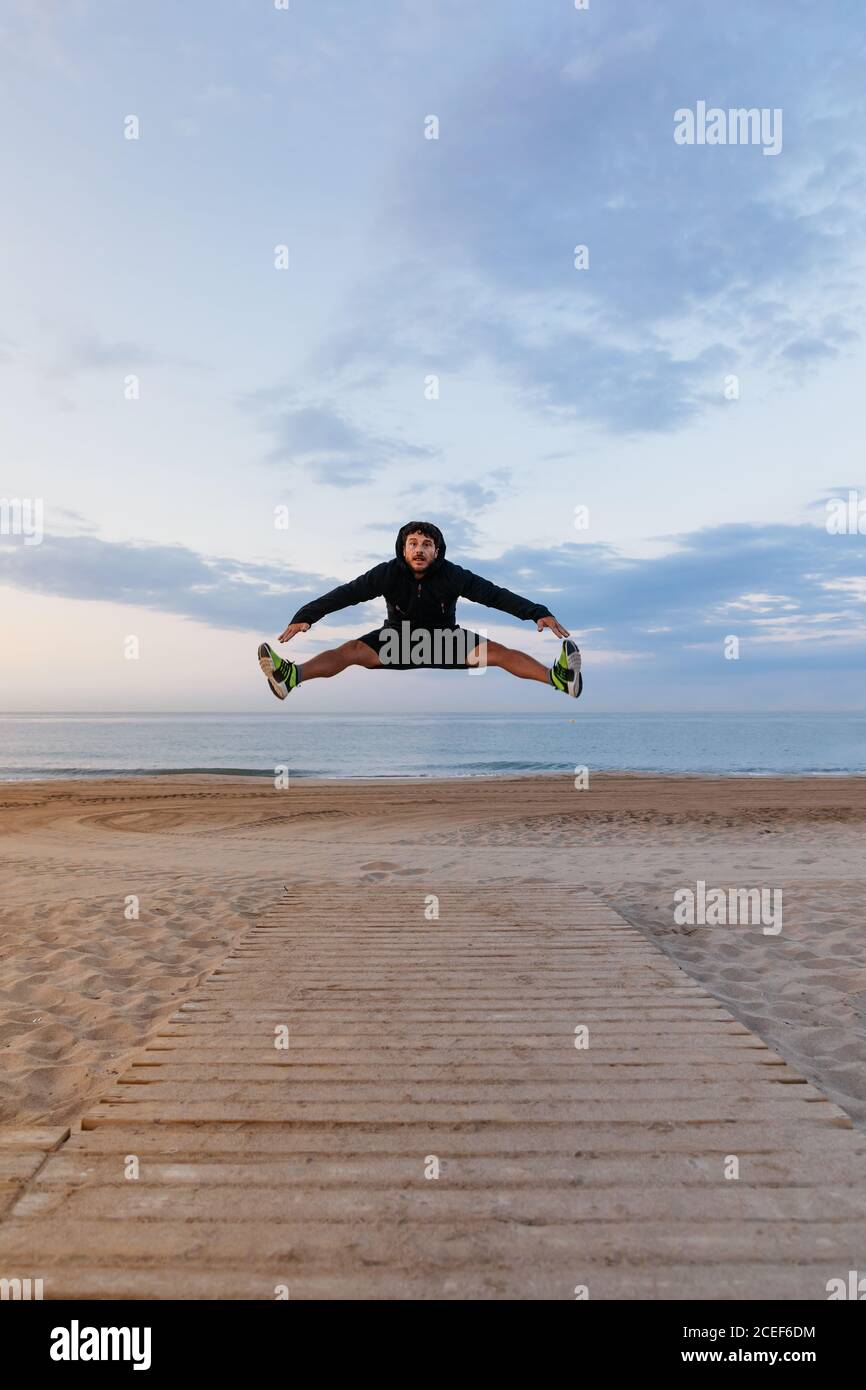 Handsome man in sportswear jumping high during outdoor training on sandy beach Stock Photo