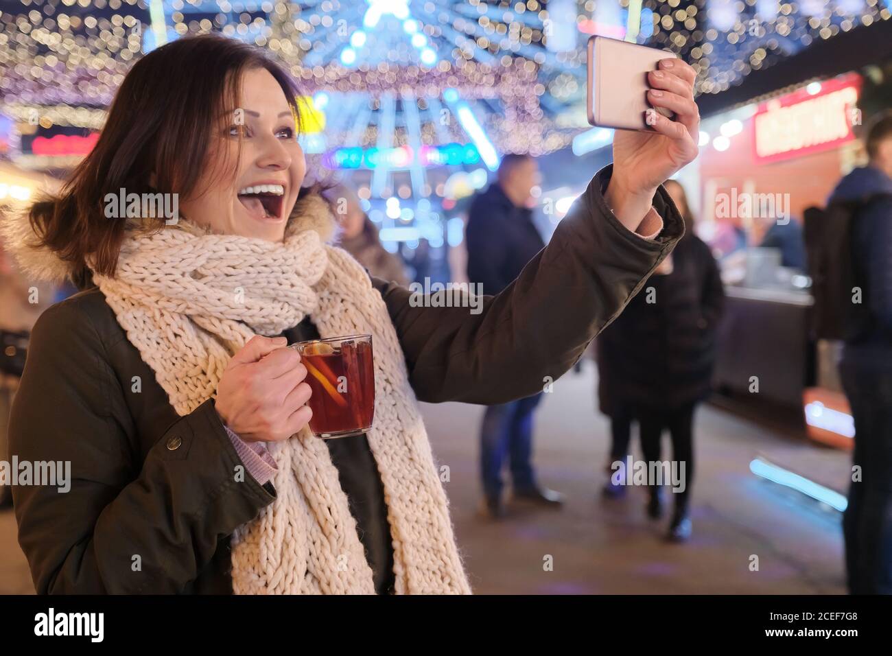 Christmas market, mature happy woman with mug of mulled wine and smartphone taking selfie Stock Photo