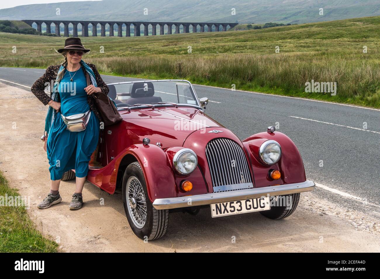A woman in a turquoise dress standing by a 2003 model  Morgan 4/4 drop-head coupé sports car, Ribblehead Viaduct, North Yorkshire, England Stock Photo