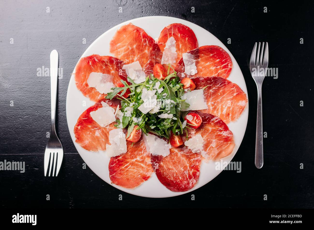 Tasty thin slices of jerked meat sprinkled with chopped tomatoes and potherbs set on white ceramic plate with forks around on black wooden table from above Stock Photo