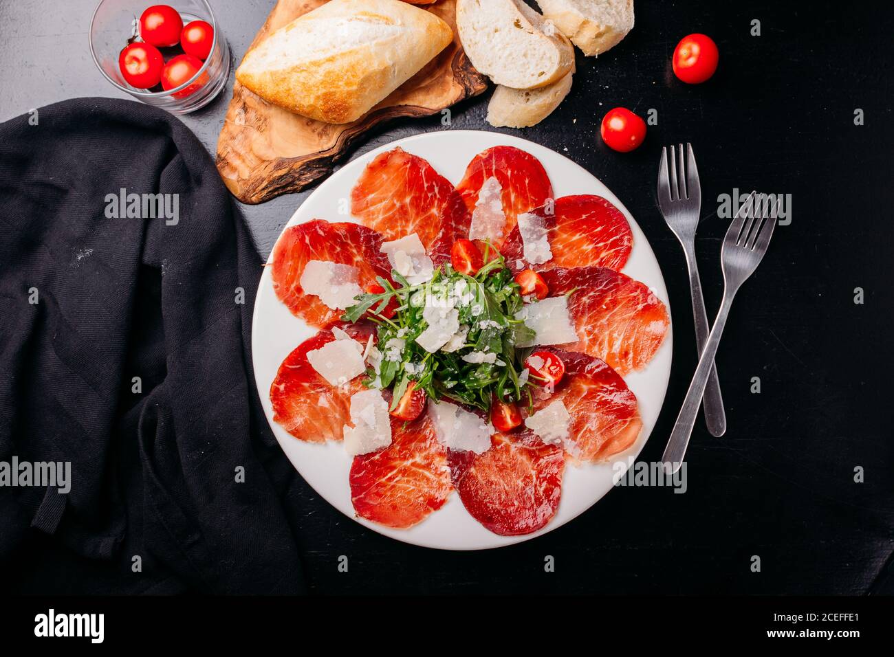 Tasty thin slices of jerked meat sprinkled with chopped tomatoes and potherbs set on white ceramic plate with forks around on black wooden table from above Stock Photo