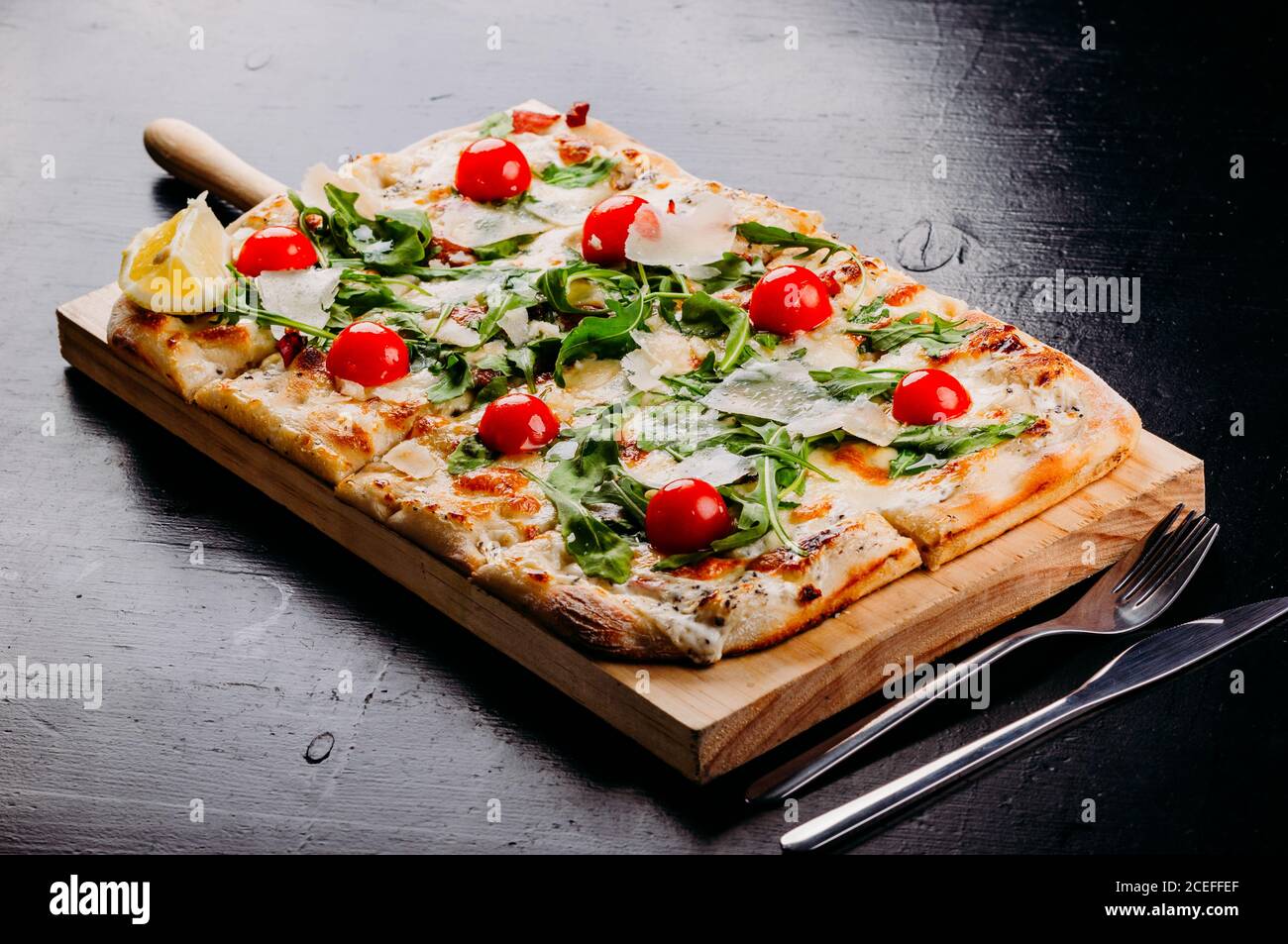 Served on wooden board fresh rectangular cut pizza with cherry tomatoes, potherbs and cheese placed on dark table with scattered flour, vegetables, fork and pizza cutter from above Stock Photo