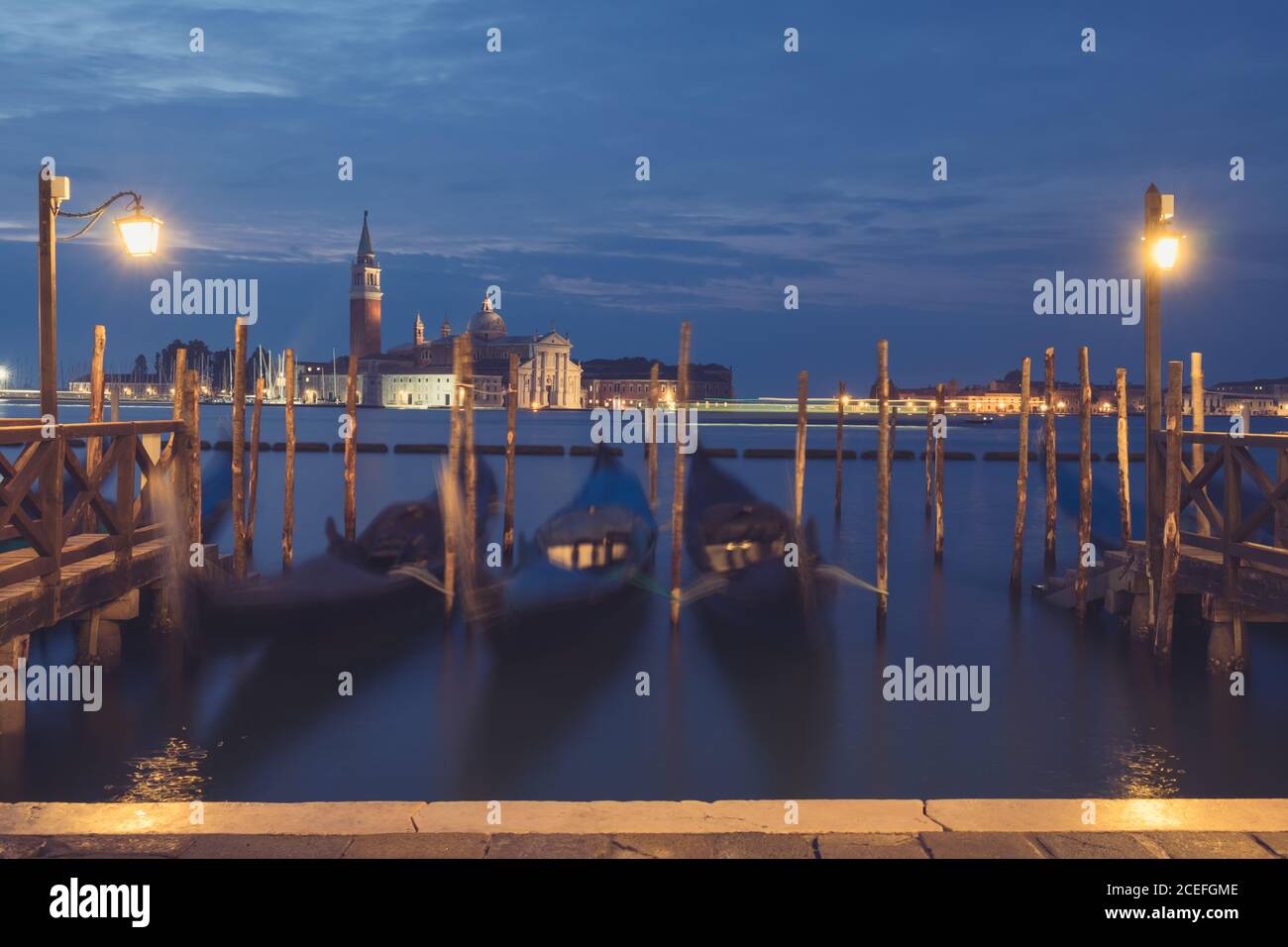 Three empty gondolas attached to wooden poles on calm canal water in