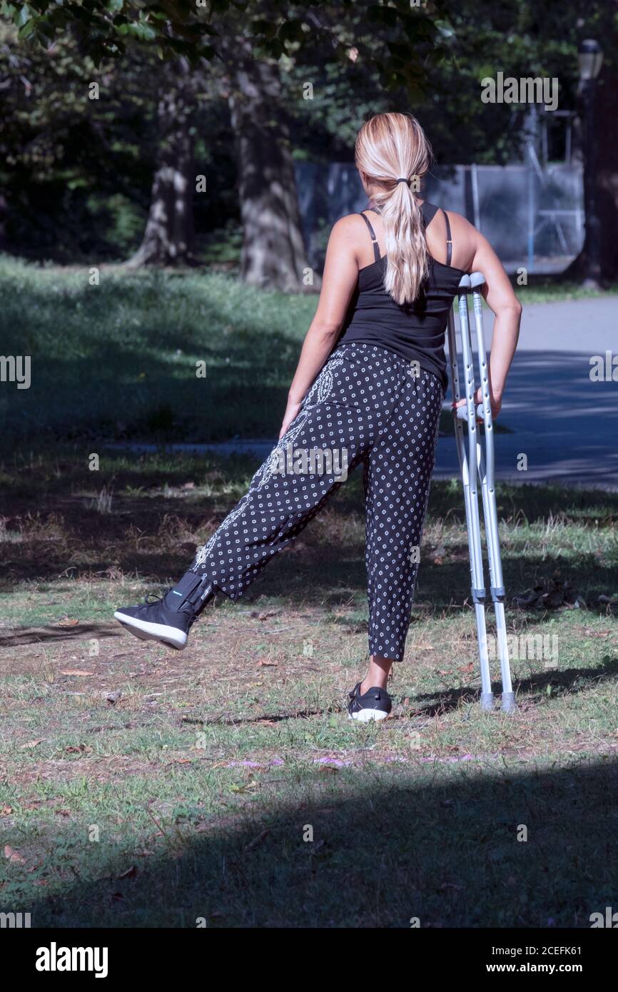 A young lady with an ankle injury stretches her other leg while leaning on crutches. In a park in Queens, New York City. Stock Photo