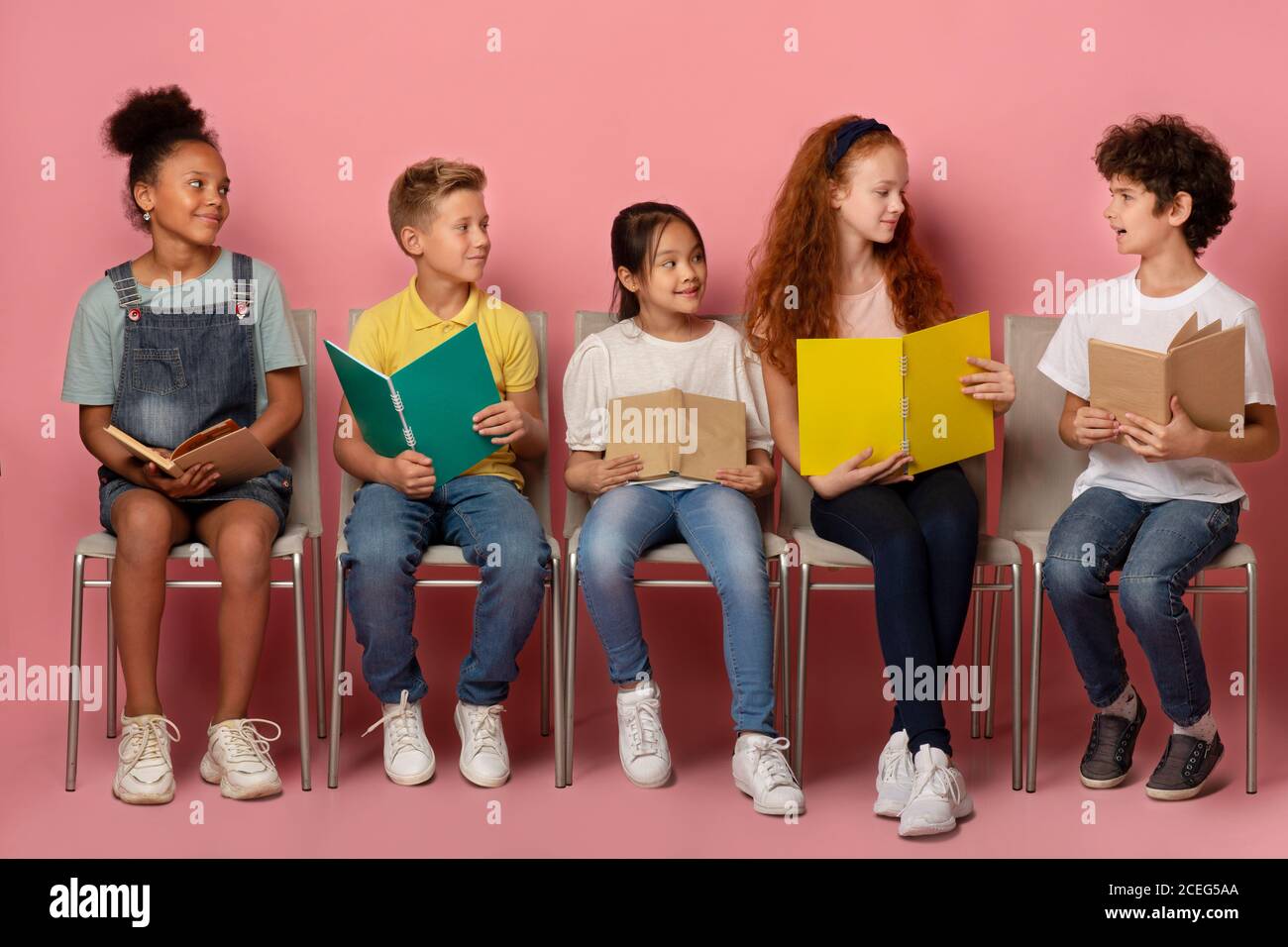 Multiracial schoolkids discussing their home assignment while sitting on chairs against pink background Stock Photo