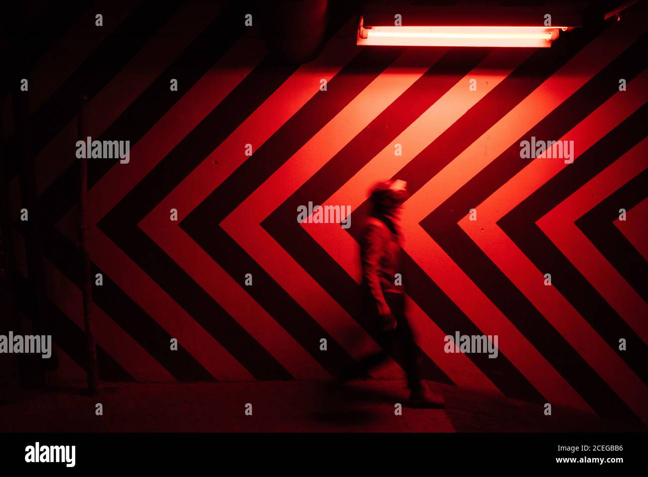 Side view of motion blurred figure of male walking down in tunnel in direction opposite to large red and black arrows on wall lit by red lamps Stock Photo