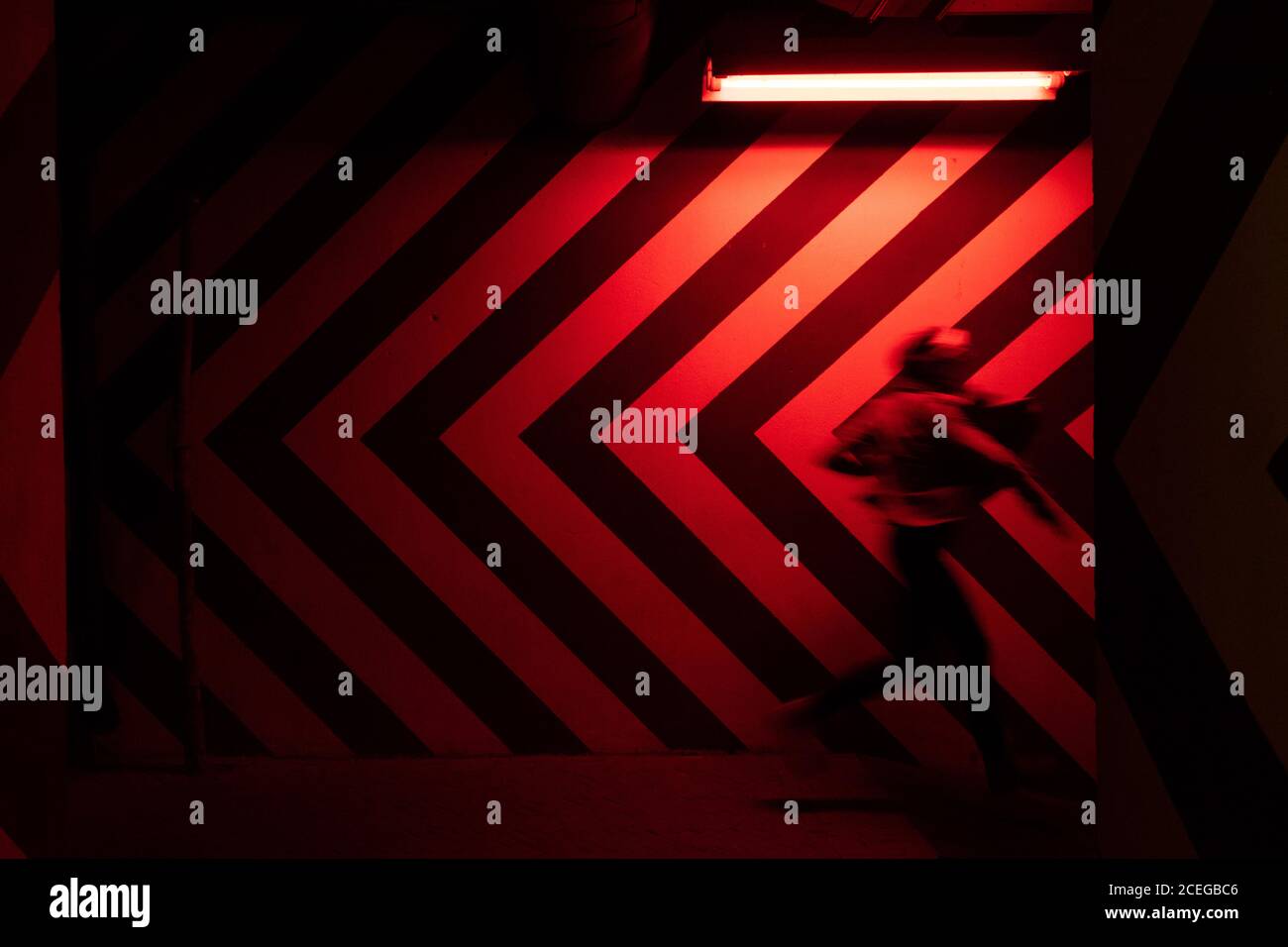 Side view of motion blurred figure of male walking down in tunnel in direction opposite to large red and black arrows on wall lit by red lamps Stock Photo