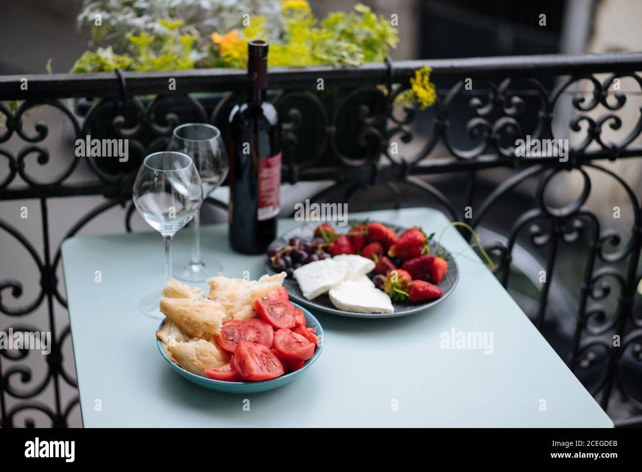 Bottle of wine and delicate glasses with high stems standing on table with sliced tomatoes, bread, cheese and berries lying on plates with ornamental fence and plants on background Stock Photo