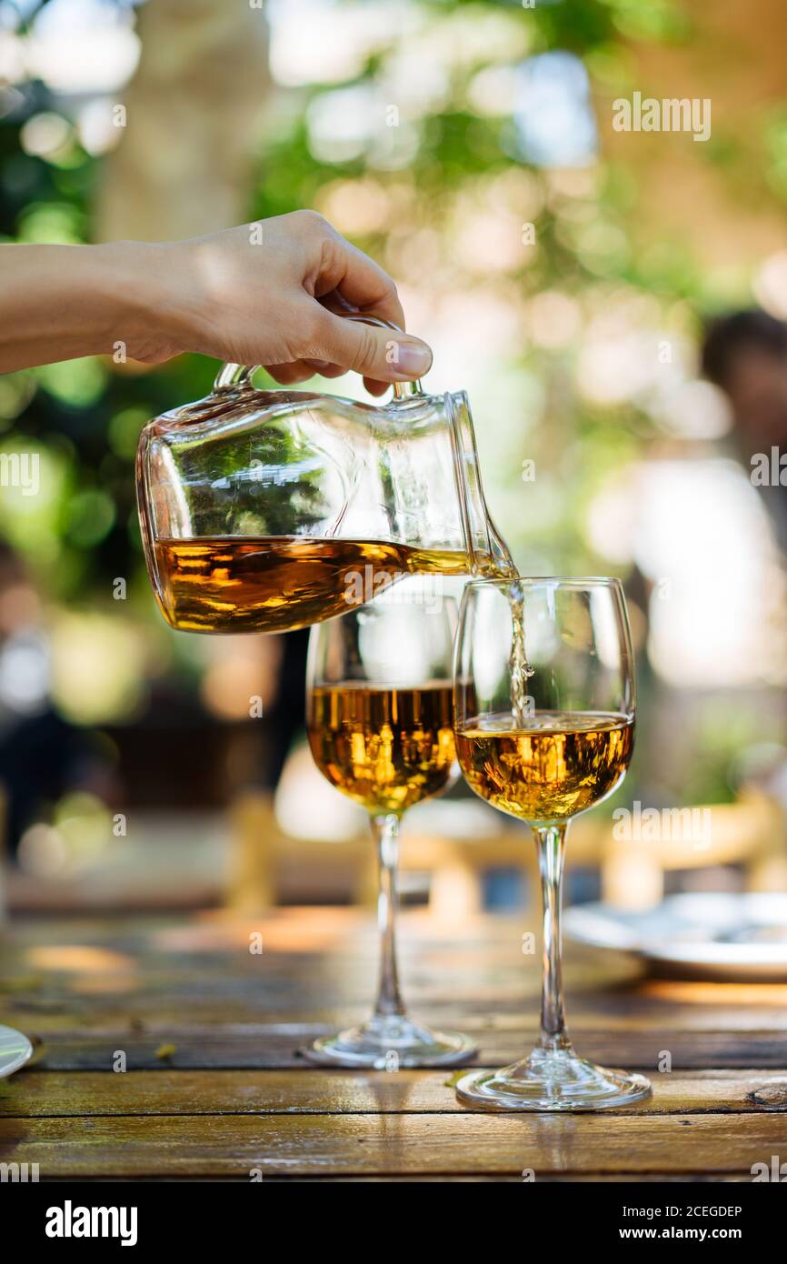 Crop hand pouring white wine from glass pitcher into glasses with high