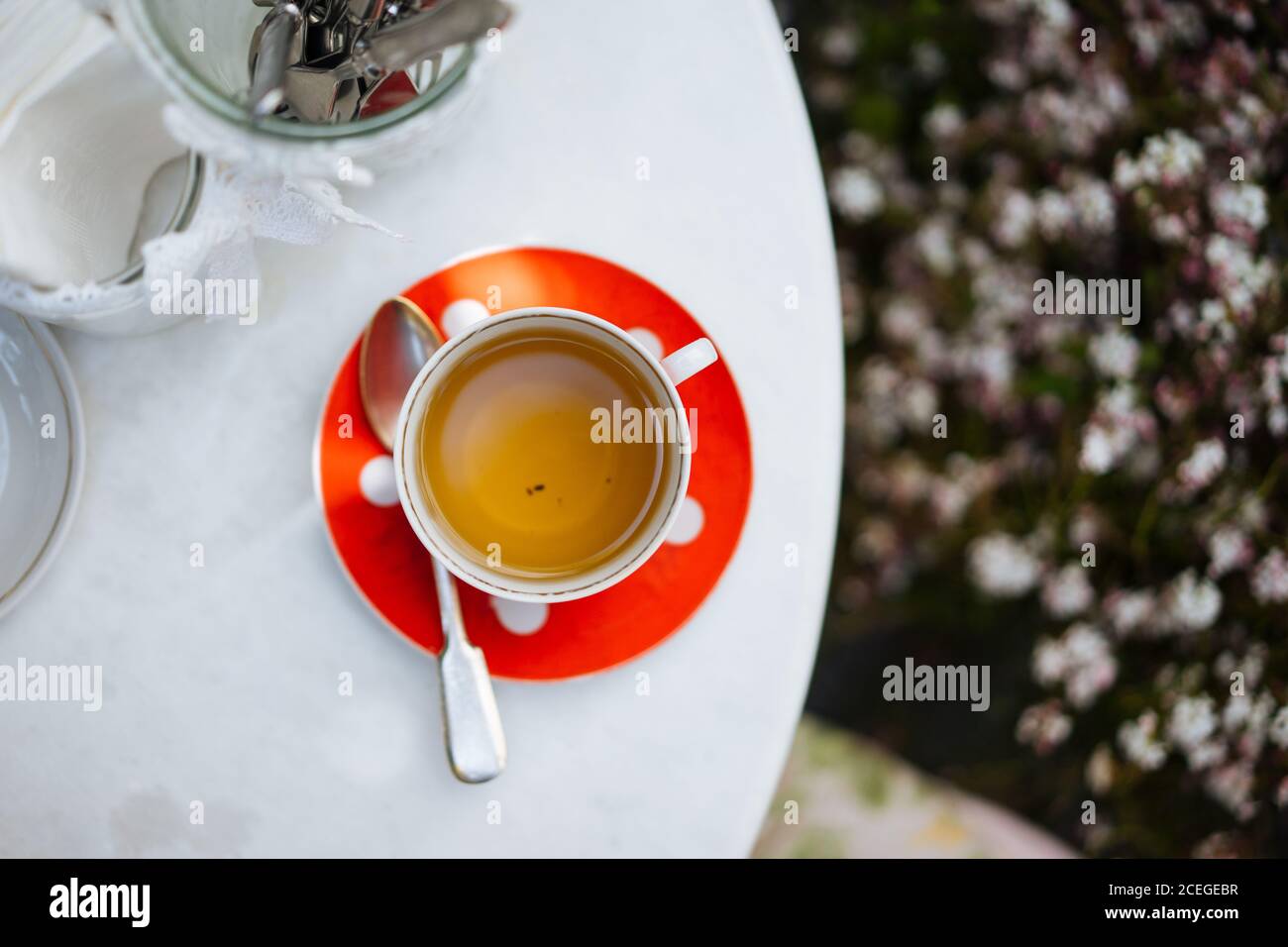 Red ceramic polka-dotted mug on saucer with spoon?and light drink in it standing on white round table with elegant jars with lace ribbons outdoors from above Stock Photo