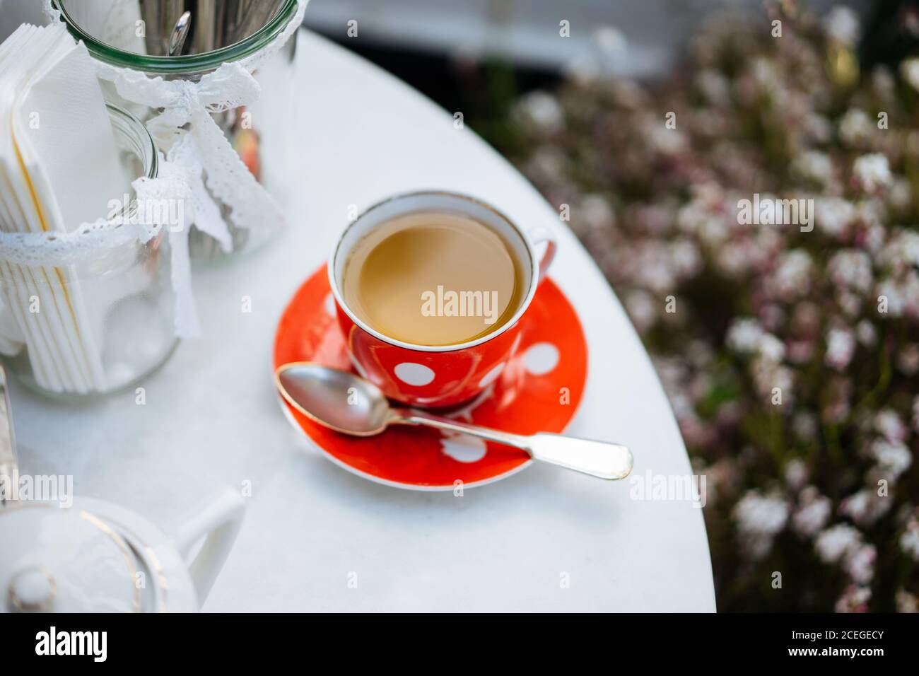 Red ceramic polka-dotted mug on saucer with spoon?and light drink in it standing on white round table with elegant jars with lace ribbons outdoors from above Stock Photo