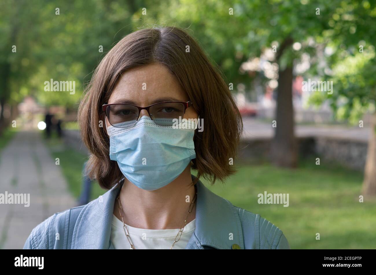 Young woman in medical mask standing in city park looking to camera green woods on background Concept of health and safety life COVID-19 coronavirus v Stock Photo