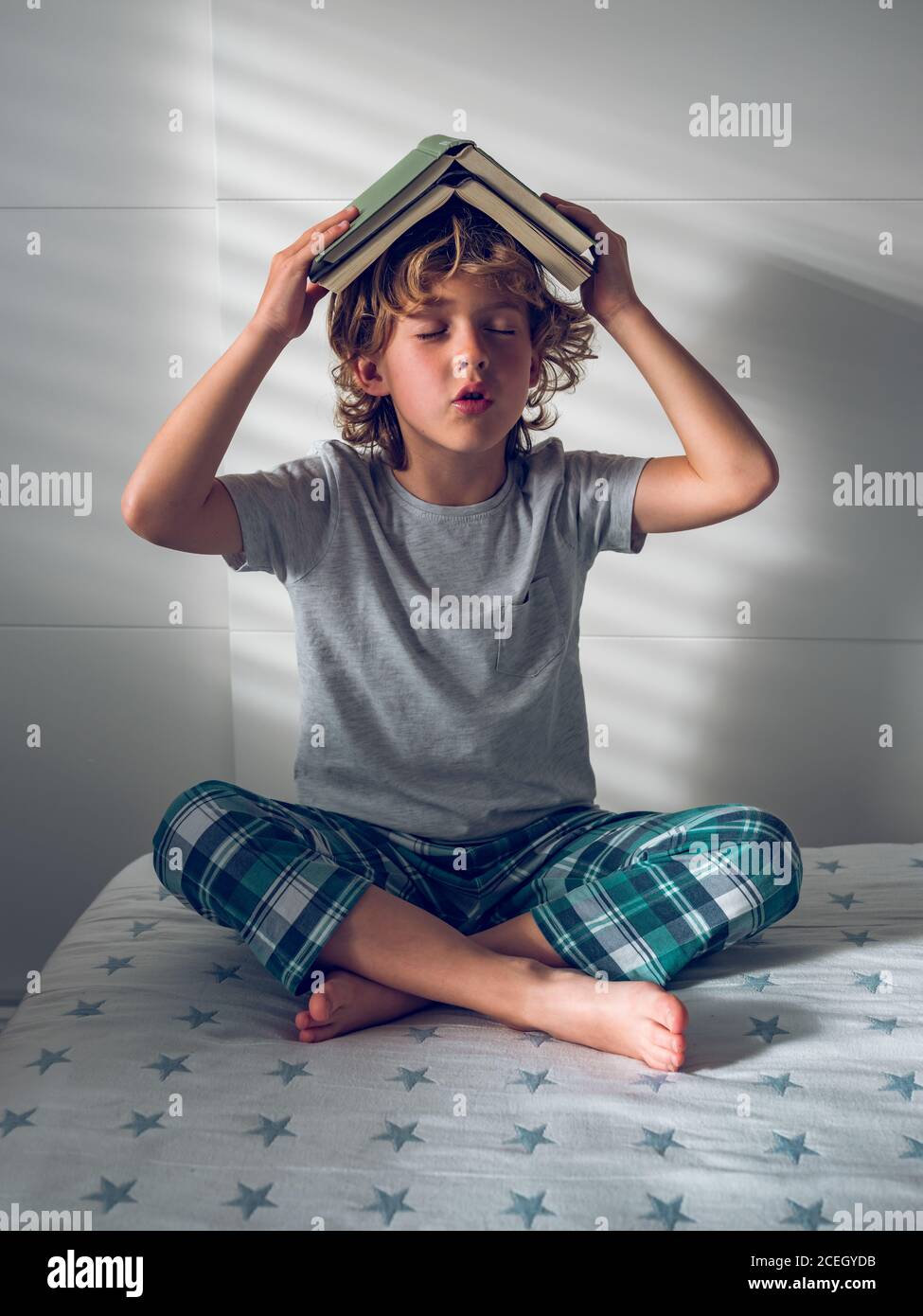 Adorable boy in pajamas wearing book on head and enjoying reading while sitting on comfortable bed Stock Photo Alamy
