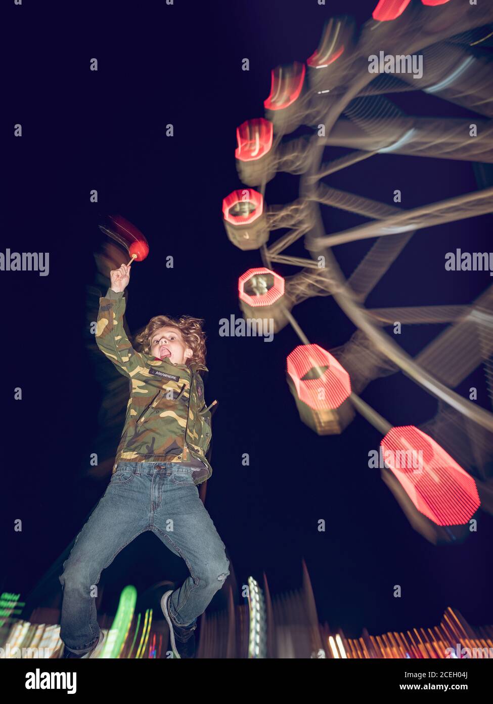 Cheerful little boy with apple jumping on the background of carousel at night. Stock Photo