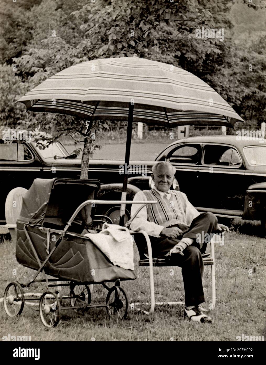 Grandfather sitting by a baby carriage under an umbrella in an urban park, circa 1947. Stock Photo