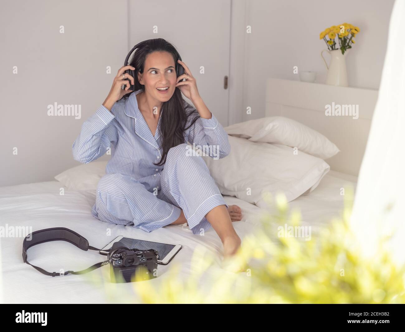 Beautiful Woman wearing blue pajamas and headphones sitting on bed,enjoying music and looking away Stock Photo