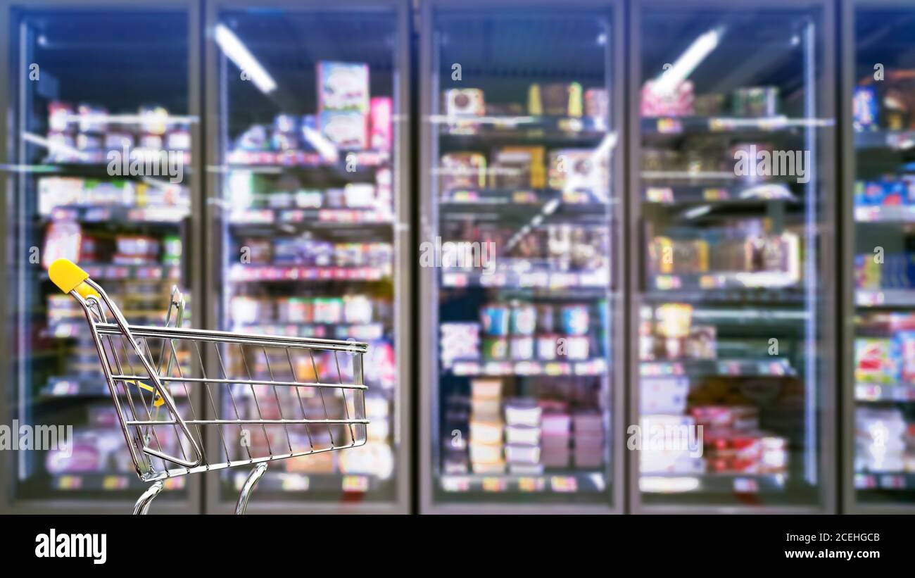 An empty shopping cart is standing in the supermarket. In the background you can see a refrigerator shelf, it is blurred and has a lot of colorful bok Stock Photo