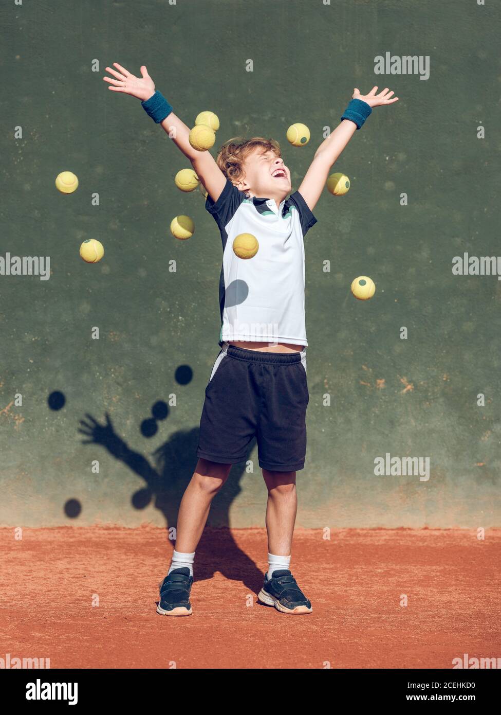 Cheerful little child with upping hands and closed eyes between suspended tennis balls in air on court in sunny day Stock Photo