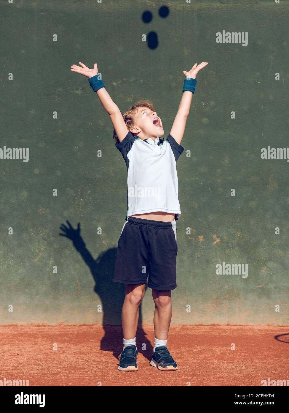 Cheerful little child with upping hands and closed eyes between suspended tennis balls in air on court in sunny day Stock Photo
