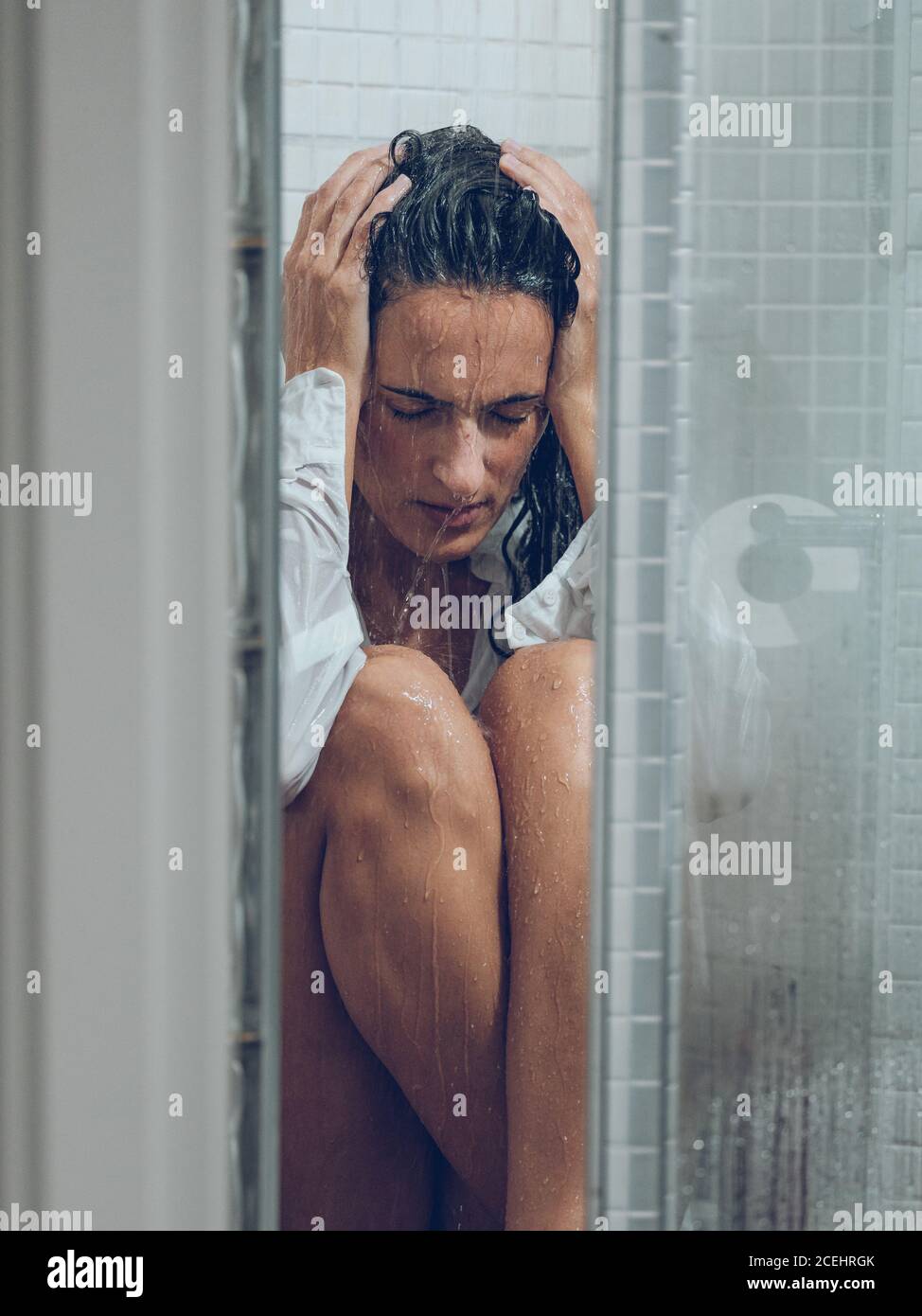 Sad young lady in white wet shirt with hands on head sitting under water spray in shower cabin Stock Photo