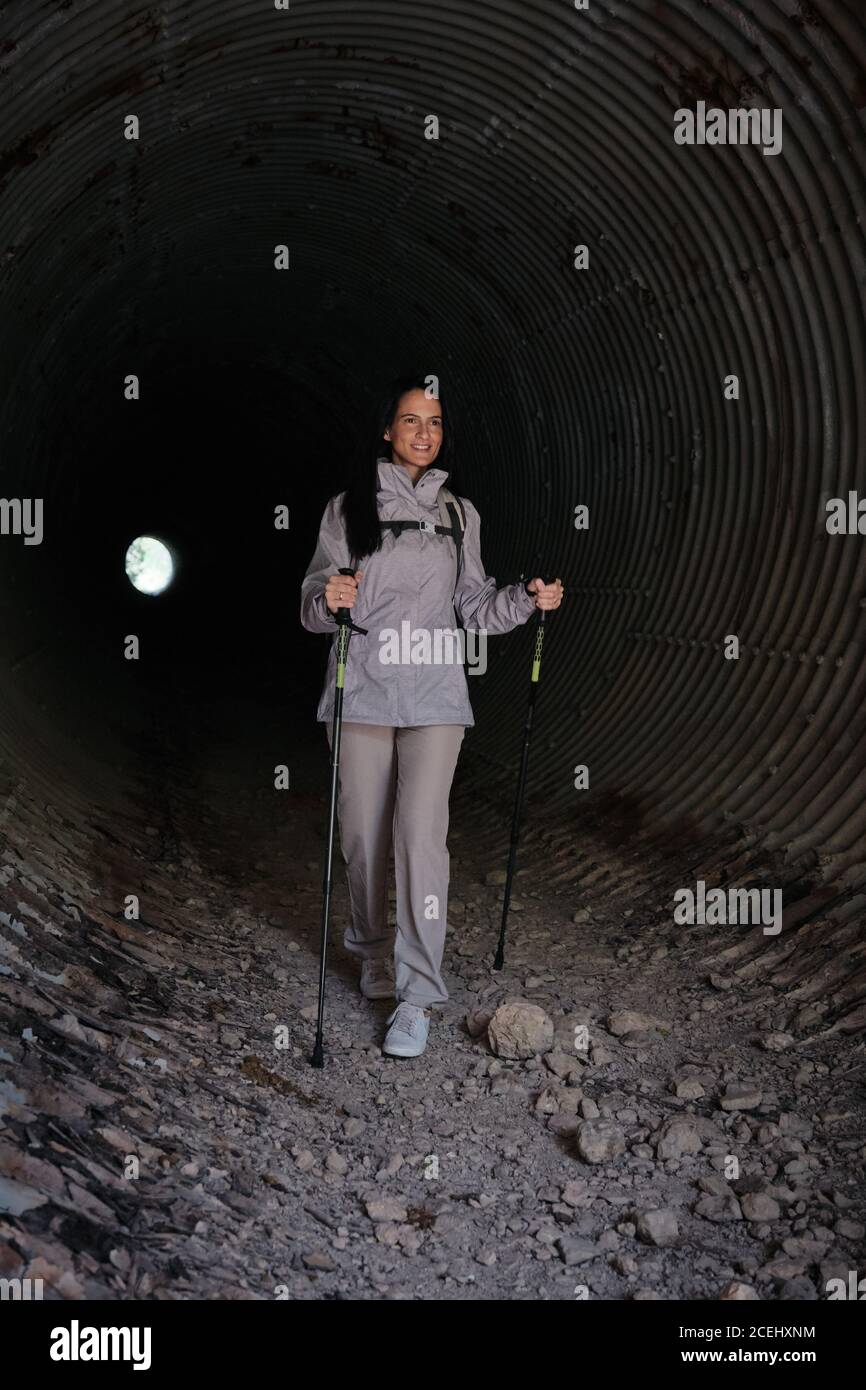 Cheerful lady in coat walking with trekking sticks in big tunnel in obscurity Stock Photo