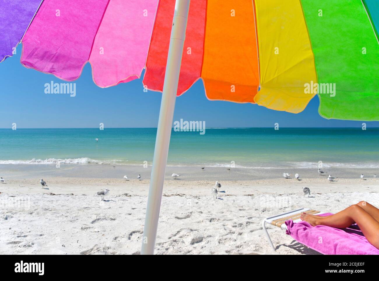 A Multi-Clored Beach Umbrella with a Woman's Legs Showing on a Chair on the Beach While Enjoying the Sunny Day. Stock Photo