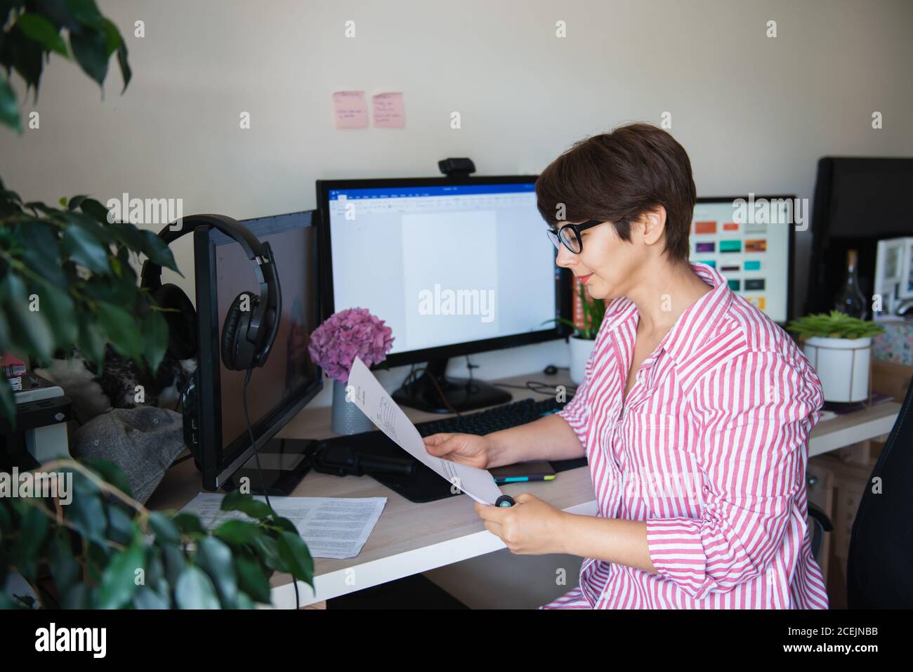 Side view woman in glasses and striped shirt reading document sitting at her work desk with few monitors at home working space. Remote work concept Stock Photo