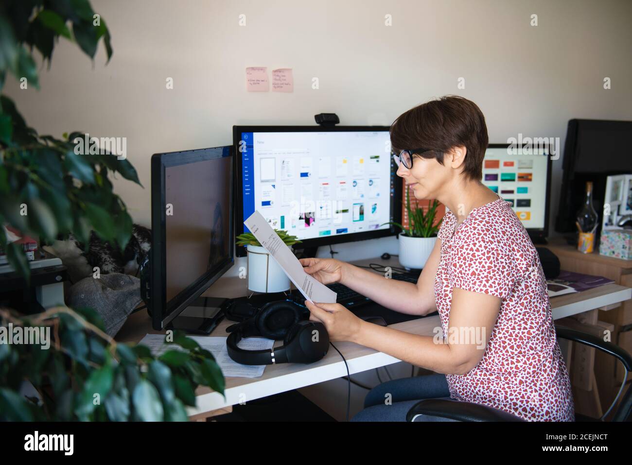 Side view woman in glasses and casual clothes reading document sitting at her work desk with few monitors at home working space. Remote work concept Stock Photo