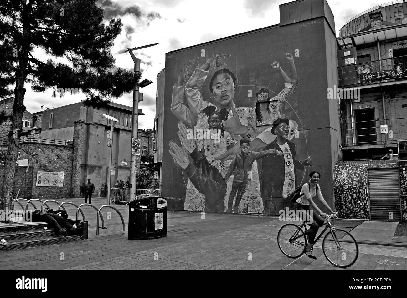 A SCENE OF GILLETT SQUARE IN DALSTON,EAST LONDON, WITH A COMMUNITY MURAL, A SMILING YOUNG WOMEN ON A BIKE & A MAN SLEEPING ON THE PAVEMENT. Stock Photo
