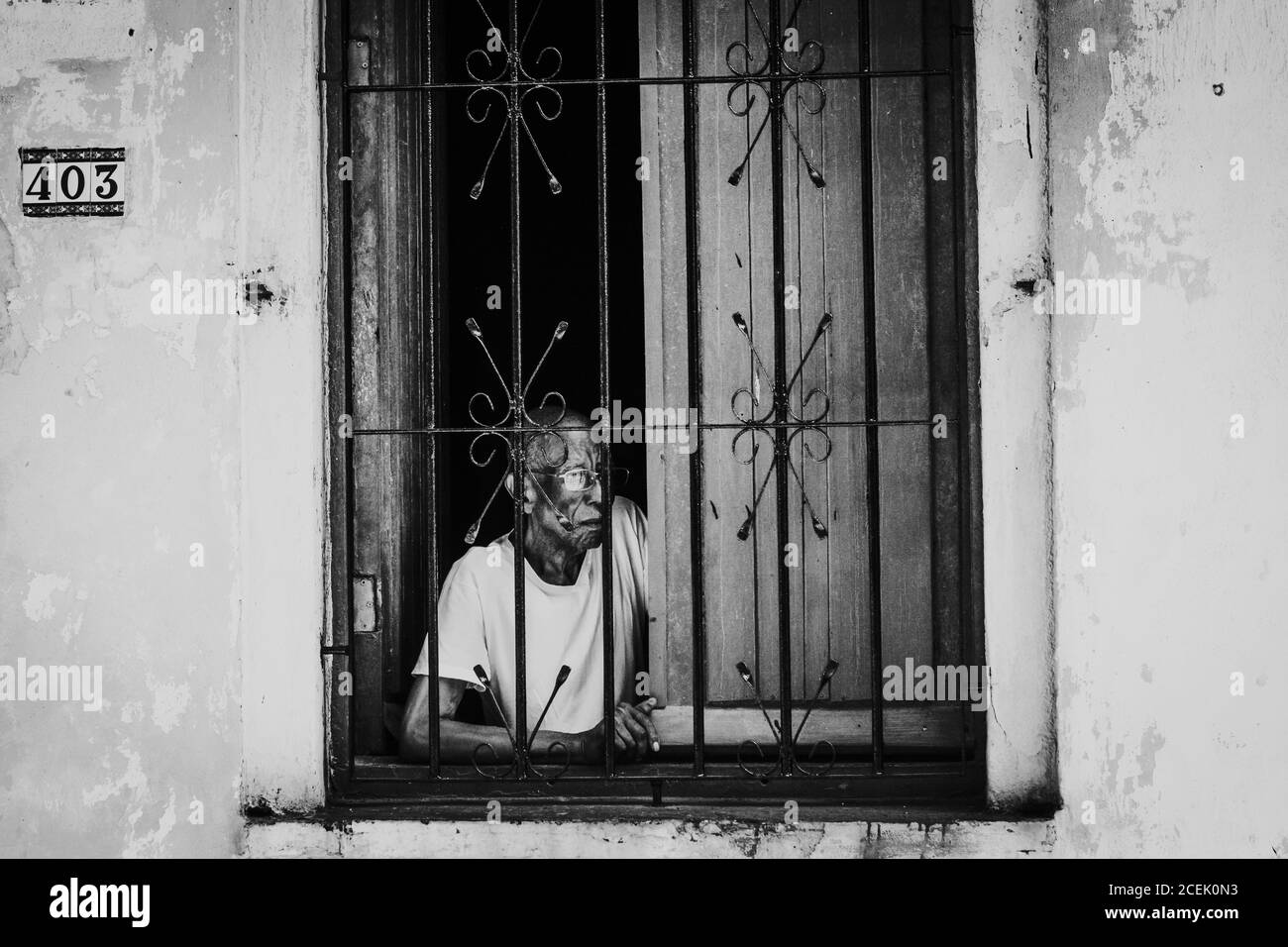LA HABANA, CUBA - MAY 1, 2018: Black and white shot of elderly man in window with metal bars looking away on street, Cuba. Stock Photo