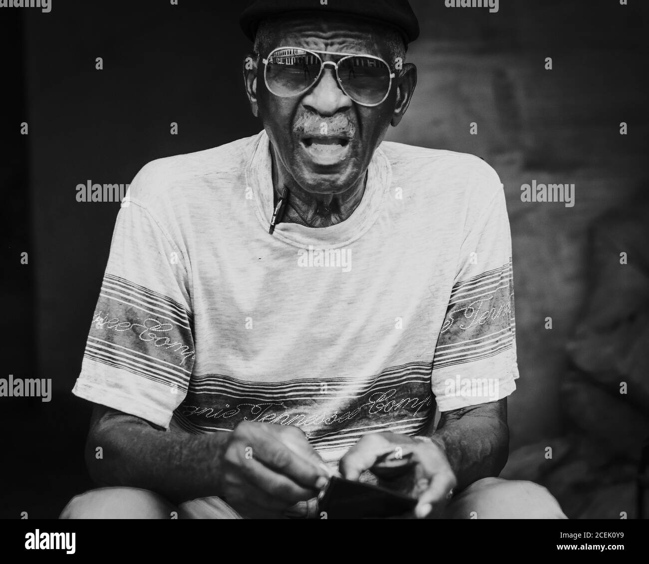 LA HABANA, CUBA - MAY 1, 2018: Elderly ethnic man in sunglasses and t-shirt with cap looking at camera sitting on street of Cuba. Stock Photo